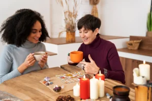 שתי נשים מחייכות בשיחת ליווי רגשי עם פתיחה בקלפי טארוט, מוקפות נרות ואבני חן
